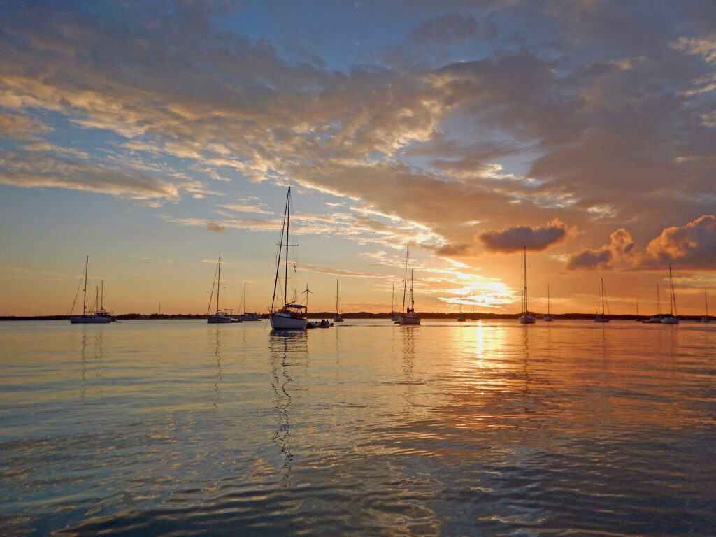 A wide bay of rippling water reflecting a beautiful sunset divided in blue and orange and the tall masts of several sailboats peacefully at anchor under an arch of clouds spreading to the horizon.